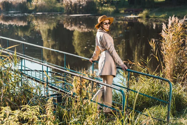 サングラス トレンチ コートと帽子ポーズを公園の池の近くで若い上品な女性  — 無料ストックフォト