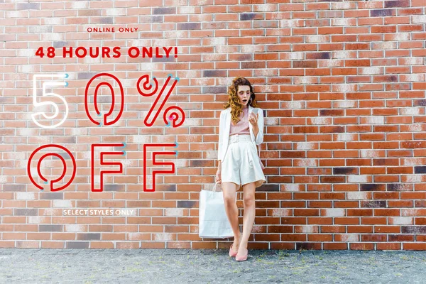 shocked young woman with shopping bags using smartphone in front of brick wall, sale banner