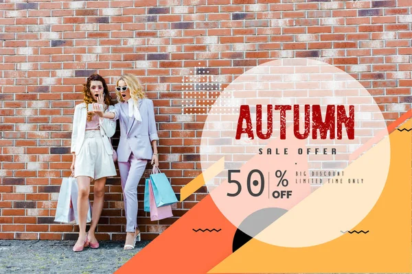 shocked young women with shopping bags taking selfie while standing in front of brick wall, autumn sale banner