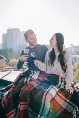 happy young couple covering in plaid and clinking glasses of red wine on rooftop clipart