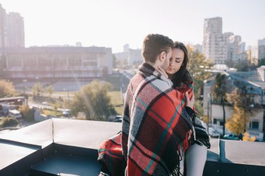 beautiful young couple covering in plaid and cuddling on rooftop clipart