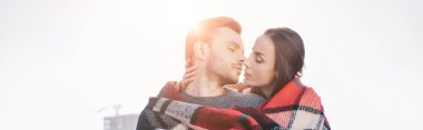panoramic shot of beautiful young couple covering in plaid and kissing with sun shining behind