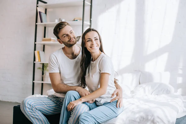 朝のベッドの上に座って よそ見はパジャマ姿で幸せな若いカップル  — 無料ストックフォト