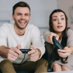 Jovem casal emocional com copos de vinho jogando jogo de vídeo retro no sofá em casa