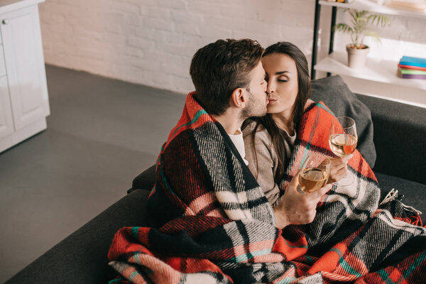 high angle view of young couple with glasses of white wine relaxing and kissing on couch under plaid
