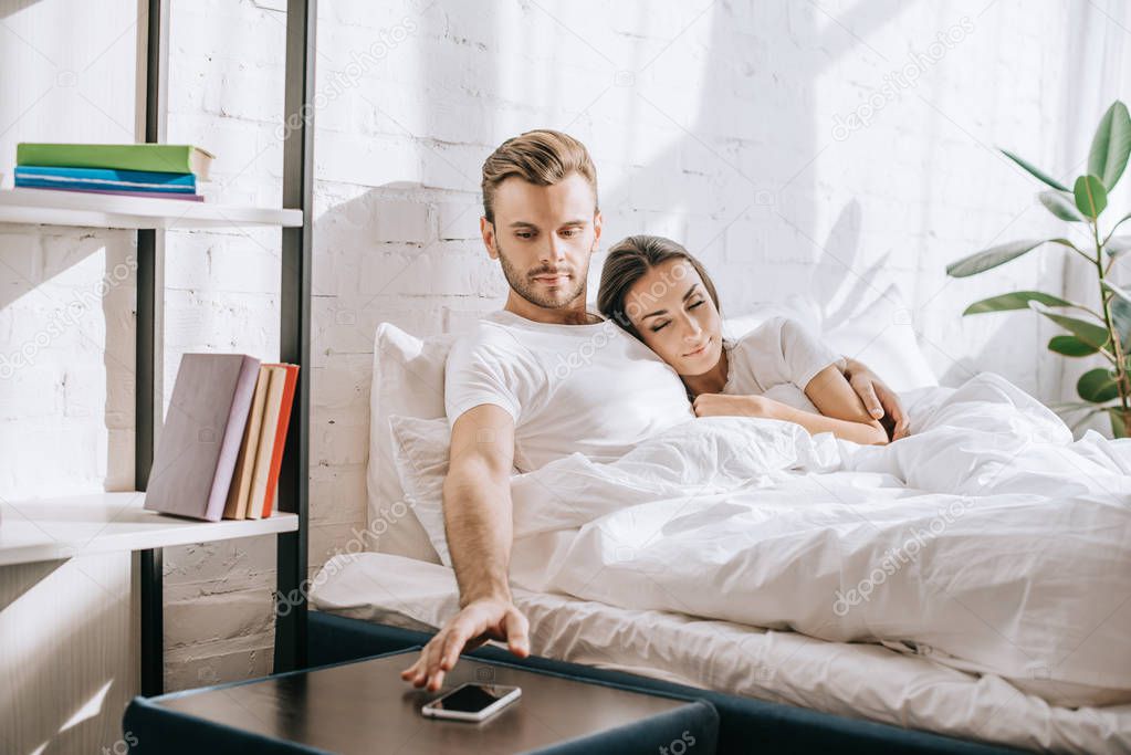 handsome young man relaxing in bed with girlfriend and reaching for smartphone