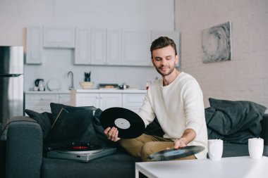 attractive young man holding discs for vinyl record player while sitting on couch clipart