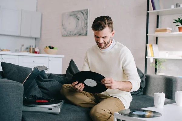 英俊的年轻人拿着光盘的乙烯基唱片播放器 而坐在沙发上 — 图库照片
