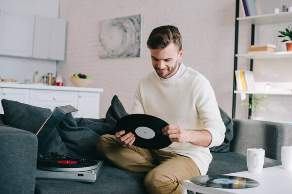 handsome young man holding disc for vinyl record player while sitting on couch