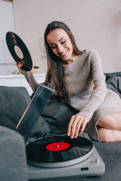 smiling young woman listening music with vinyl record player on couch at home