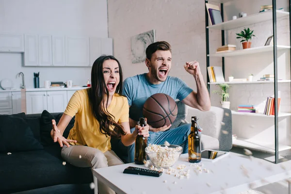 富有表现力的年轻夫妇与啤酒欢呼的篮球比赛在家里 — 图库照片
