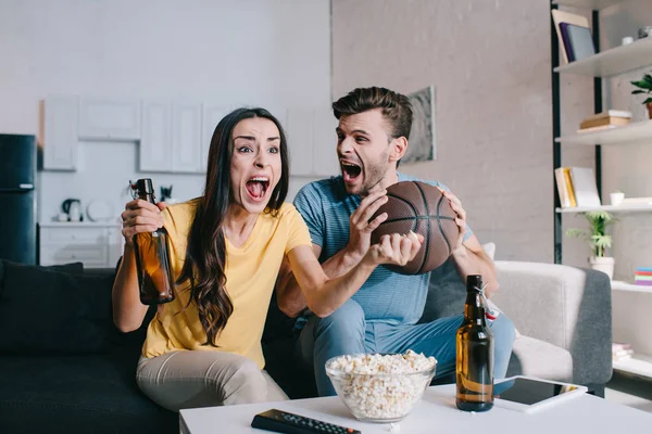 在家里为篮球比赛欢呼的年轻夫妇喝啤酒 — 图库照片