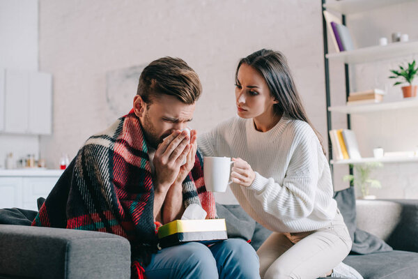 beautiful young woman holding cup of warming drink for sick boyfriend while he sneezing