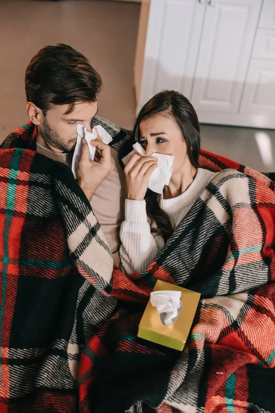 ill young couple with paper napkins sneezing while sitting on couch under plaid at home