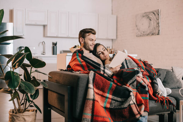 smiling young couple reading book together on couch under plaid at home