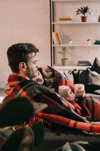 beautiful young couple with cups of cocoa with marshmallow relaxing on couch under plaid at home