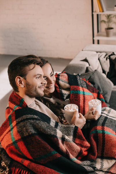 smiling young couple with cups of cocoa with marshmallow relaxing on couch under plaid at home