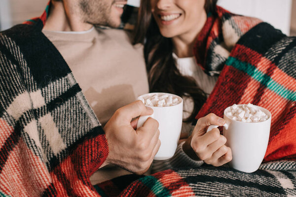 cropped shot of smiling young couple with cups of cocoa with marshmallow relaxing on couch under plaid at home