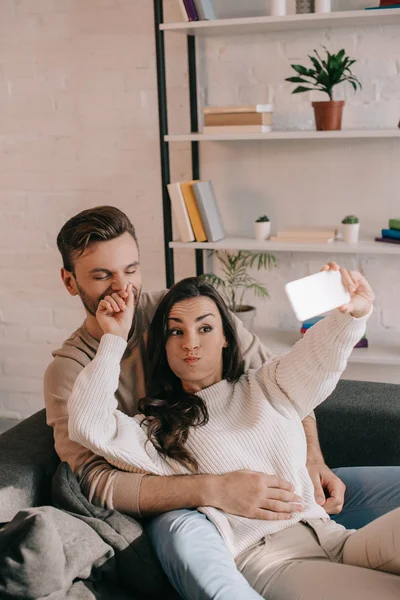 自宅のソファでスマート フォンで Selfie を取って遊び心のある若いカップル  — 無料ストックフォト