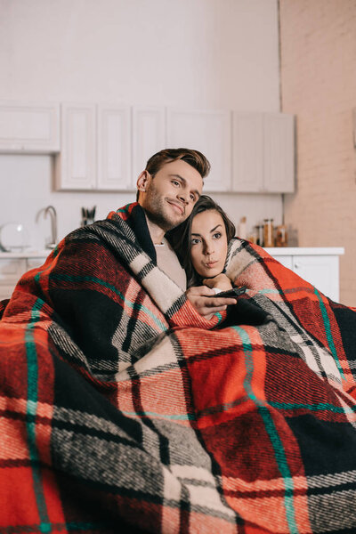 emotional young couple relaxing on couch and watching tv together while covering with plaid