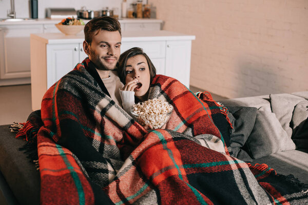 beautiful young couple watching movie with popcorn on couch and covering with plaid