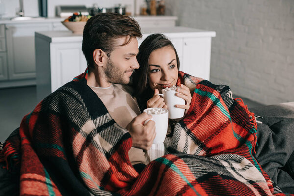 happy young couple with mugs of cocoa with marshmallow relaxing on couch and covering with plaid