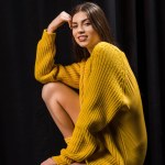 Side view of smiling woman in yellow woolen sweater on wooden bar stool on black backdrop