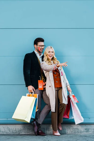 Lächelndes Stilvolles Paar Mit Einkaufstaschen Mit Verkaufsetiketten Und Posiert Blauer — kostenloses Stockfoto