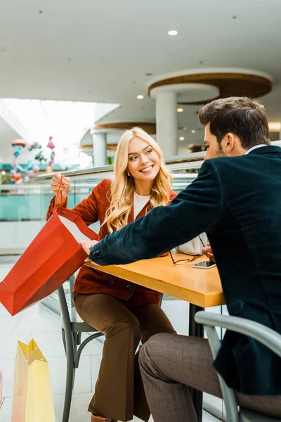 微笑的女人显示的东西在购物袋男友 而坐在咖啡馆在购物中心 — 免费的图库照片