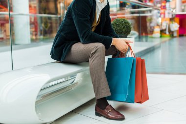 Alışveriş torbaları tutarak ve alışveriş merkezinde oturan şık adam görünümünü kırpılmış 
