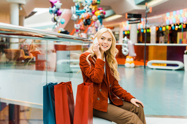 fashionable young woman talking on smartphone and sitting with bags in shopping mall