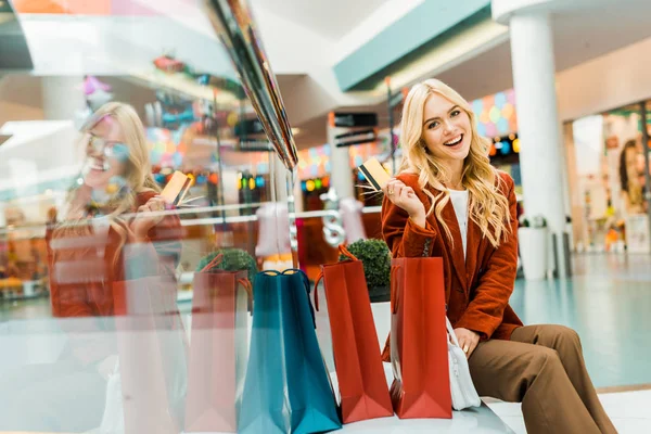 兴奋的女人拿着信用卡 坐在商场里拿着购物袋 — 图库照片