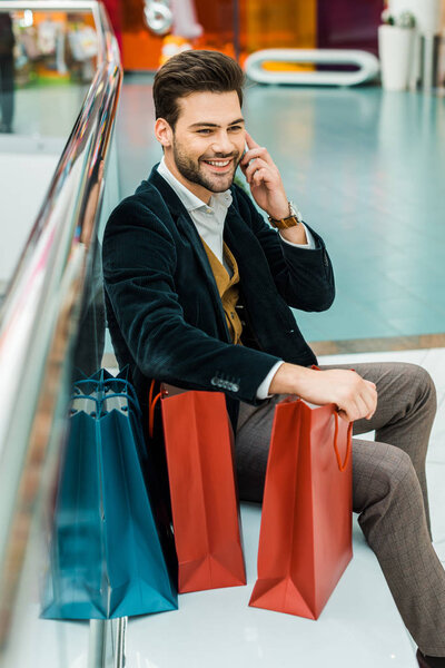 handsome smiling man talking on smartphone while sitting in shopping mall with bags