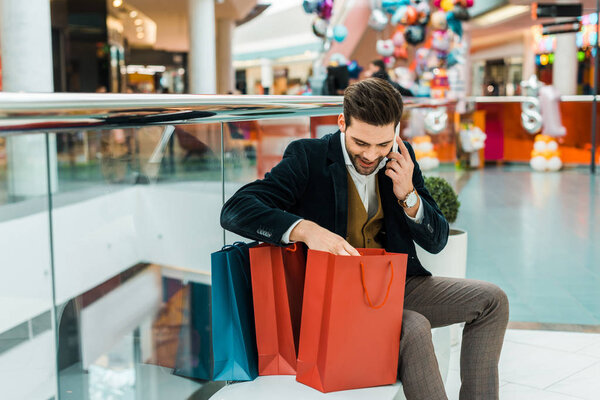 handsome man talking on smartphone and looking into bags while sitting in shopping mall 