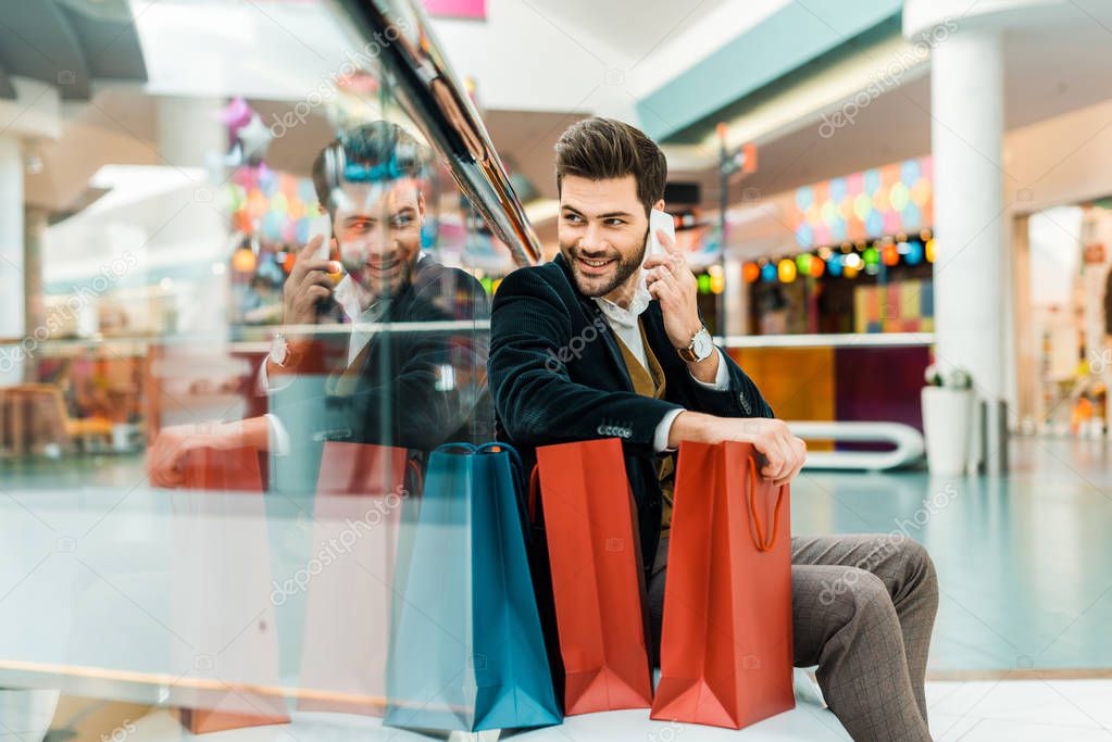 smiling man talking on smartphone while sitting in shopping mall with bags