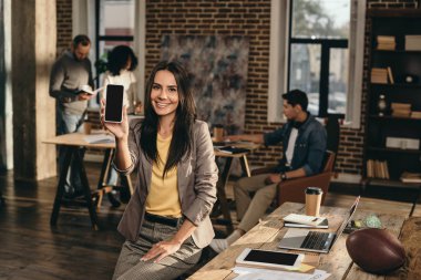 smiling casual businesswoman holding smartphone with blank screen in loft office with colleagues working behind clipart