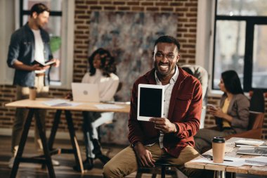 Afro-Amerikan rahat işadamı holding tablet boş ekran ve arkasında çalışma çatı office meslektaşları ile gülümseyen