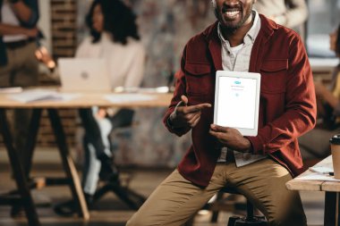 Afro-Amerikan rahat işadamı holding tablet skype ile gülümseyen görünümü kırpılmış app ve arkasında çalışma arkadaşları çatı office