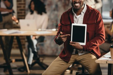Afro-Amerikan rahat işadamı holding tablet boş ekran ve arkasında çalışma çatı office meslektaşları ile gülümseyen kırpılmış görünümü