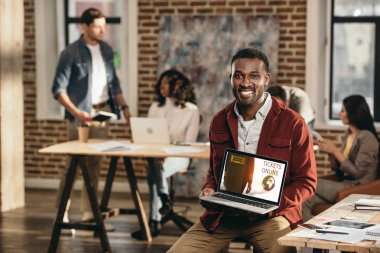 Afro-Amerikan rahat işadamı dizüstü online bilet web sitesi ve arkasında loft ofiste çalışma arkadaşları ile tutarak