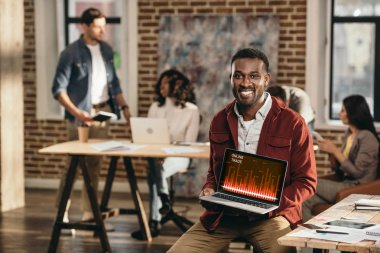 Laptop ile grafik ekran ve arkasında çalışma çatı office meslektaşları üzerinde tutarak Afro-Amerikan rahat iş adamı