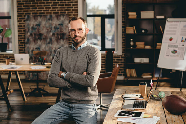 serious casual businessman sitting at desk with arms crossed in loft office