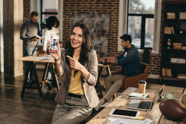微笑的休闲女商人拿着智能手机与 Pinterest 应用程序在阁楼办公室与同事工作的背后 — 图库照片