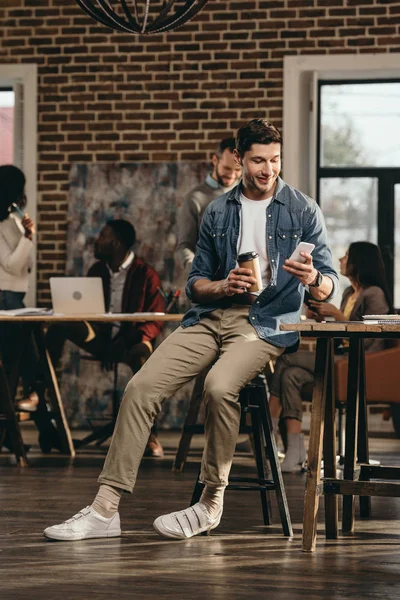 若い男が一杯のコーヒーを保持していると 背景に同僚とモダンなロフト オフィスでスマート フォンを使用して — ストック写真