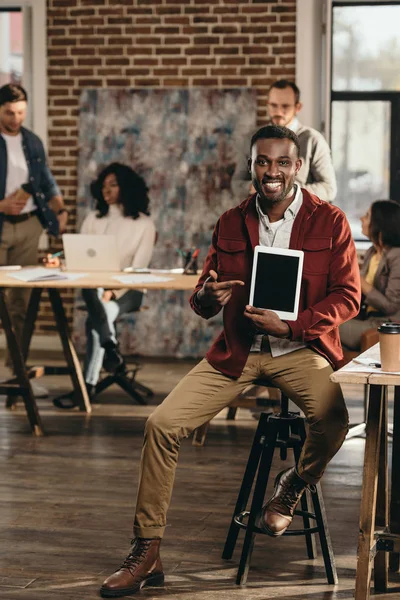 Afro-Amerikan rahat işadamı holding tablet boş ekran ve arkasında çalışma çatı office meslektaşları ile gülümseyen