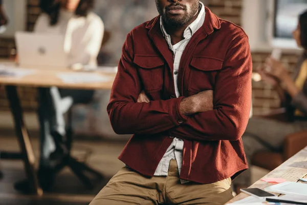 腕を組んで座っているカジュアルのアフリカ系アメリカ人のビジネスマンとの背後にあるロフトのオフィスで作業している同僚のビューをトリミング — ストック写真