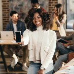 Souriant afro-américaine casual femme d'affaires pointant vers smartphone avec écran blanc dans le bureau loft avec des collègues derrière