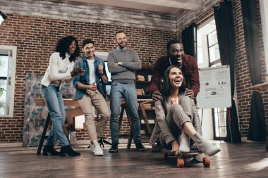 cheerful group of multiethnic coworkers having fun with skateboard in loft office clipart
