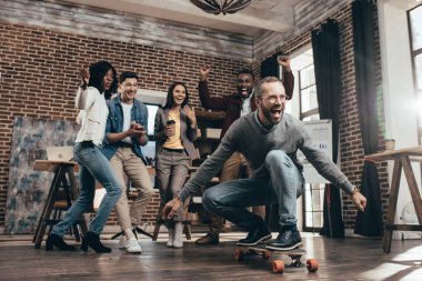 group of multiethnic coworkers having fun with skateboard in loft office clipart