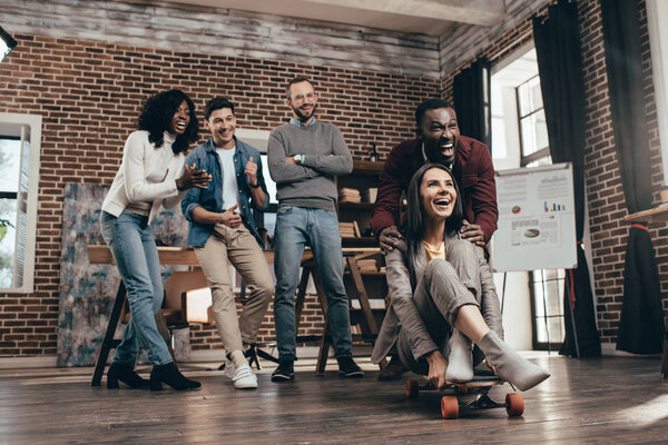 cheerful group of multiethnic coworkers having fun with skateboard in loft office